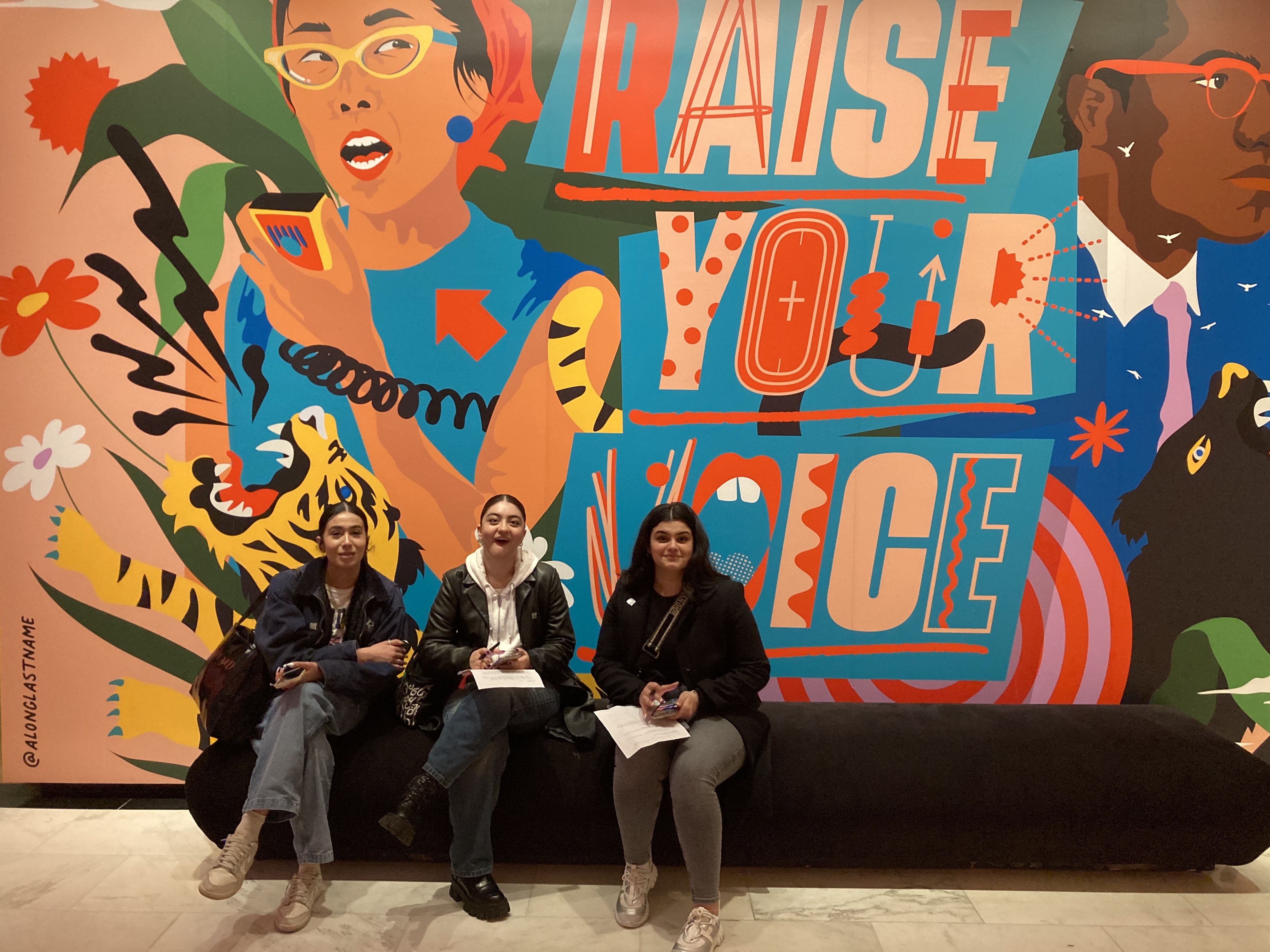 Three students sitting in front of a brightly-painted mural